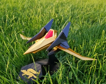 May include: A gold and black model spaceship with red accents, resting on a black stand with a gold emblem. The spaceship has two dark blue wings and is set against a backdrop of green grass.
