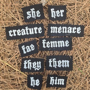 May include: A selection of black fabric patches with white gothic lettering. The patches display words and pronouns including "she", "her", "creature", "menace", "fae", "femme", "they", "them", "he", and "him". The patches are arranged on a bed of brown pine needles.