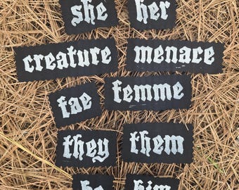 May include: A collection of black fabric patches with white gothic lettering. The patches display words and pronouns such as "she", "her", "creature", "menace", "fae", "femme", "they", "them", "he", and "him". The patches are arranged on a bed of brown pine needles.