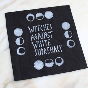 May include: Black fabric patch with white embroidered text that reads "Witches Against White Supremacy" and a circle of white embroidered moons.