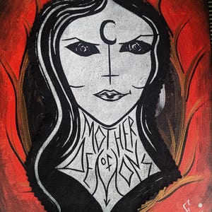 May include: A painting of a woman with long black hair and a white face. She has a crescent moon and a cross on her forehead. The text "MOTHER OF DEMONS" is written on her chest. The background is red and orange.