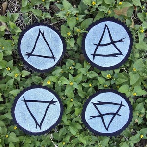 May include: Four circular patches with black trim and light blue centers, each featuring a different black geometric symbol. The patches are arranged on a bed of green leaves with small yellow flowers.