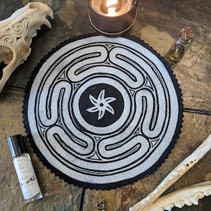 May include: A black and white circular fabric patch with a five-pointed star in the center and a repeating pattern of curved lines. The patch is surrounded by a black border and is lying on a wooden surface.