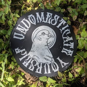 以下が含まれることがあります： 円形の黒いパッチで、白い鳥のイラストが円の中に描かれています。「UNDOMESTICATE YOURSELF」の文字が鳥の周りに様式化されたフォントで印刷されています。パッチは緑の葉の背景に配置されています。