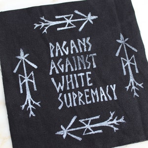 May include: Black fabric patch with white text that reads "Pagans Against White Supremacy". The patch is decorated with white symbols.