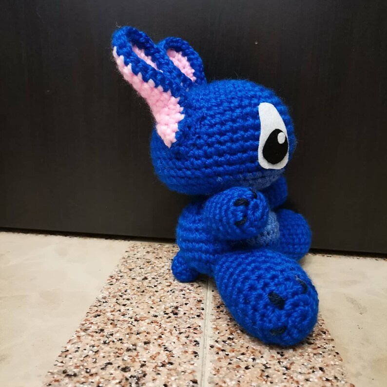 Puede incluir: Peluche de Stitch de color azul tejido a crochet con orejas rosas y un ojo blanco. El juguete est&aacute; sentado en un suelo de baldosas.