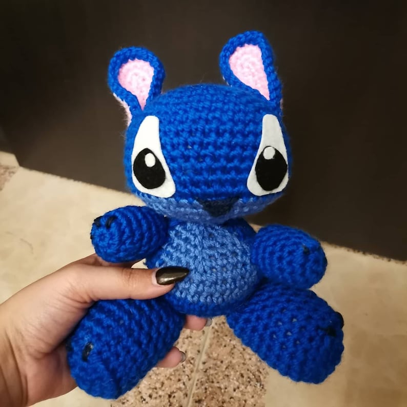 Puede incluir: Animal de peluche azul tejido a crochet con orejas rosas y ojos negros. El animal est&aacute; sentado en una mano.