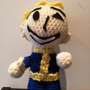 Puede incluir: Un juguete de peluche tejido a crochet de un personaje con cabello amarillo, un mono azul y un cinturón amarillo. El personaje tiene una cara blanca con ojos negros y una boca sonriente.