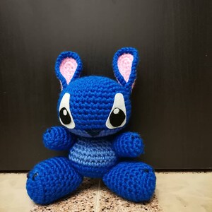 Puede incluir: Juguete de peluche azul de crochet con orejas rosas y ojos blancos. El juguete tiene forma de personaje de dibujos animados con una boca cosida.