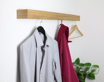 May include: A wooden wall-mounted shelf with two hangers. A gray long-sleeved shirt hangs on one hanger and a burgundy jacket hangs on the other. A wooden hanger with a pink ribbon hangs below the jacket.