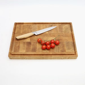 Op de afbeelding: Een houten snijplank met een lichtbruine afwerking. Een klein trosje rode tomaten en een zilveren mes liggen op de plank.