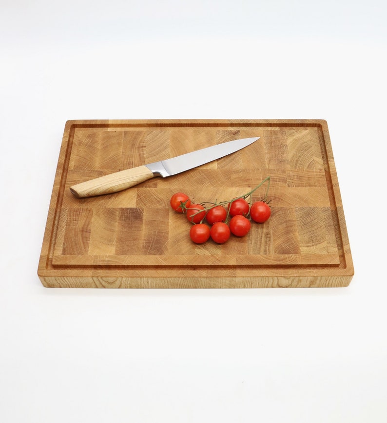 Op de afbeelding: Een houten snijplank met een lichtbruine afwerking. Een klein trosje rode tomaten en een zilveren mes liggen op de plank.