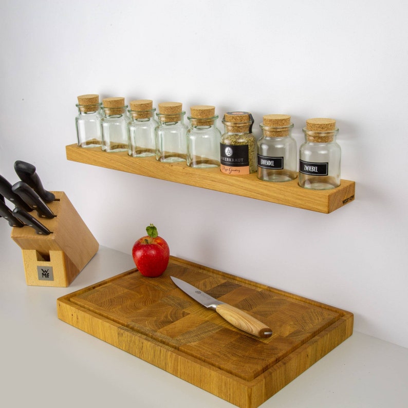 Op de afbeelding: Een houten kruidenrek met acht glazen potten met kurken deksels. De potten zijn gevuld met kruiden en gelabeld met de namen van de kruiden. Het rek is gemonteerd aan een witte muur. Een houten snijplank met een mes ligt op een wit aanrecht onder het rek.