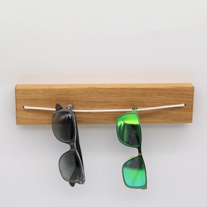 May include: A wooden wall-mounted sunglass holder with a white cord strung across it. Two pairs of sunglasses are hanging from the cord, one with black frames and dark lenses, and the other with black frames and green mirrored lenses.