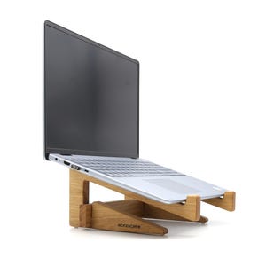 Könnte beinhalten: Ein silberner Laptop steht auf einem Holzständer. Der Ständer ist aus hellem Holz gefertigt und hat ein minimalistisches Design. Der Bildschirm des Laptops ist schwarz. Der Markenname "WOODKOPF" ist auf dem Ständer zu sehen.