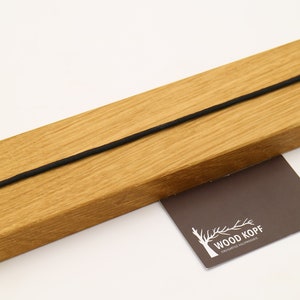 May include: A wooden wall mount with a black elastic cord running through it. The mount is made of light brown wood and has two screw holes on the back. The mount is labeled "WOOD KOPF" on a small brown card.