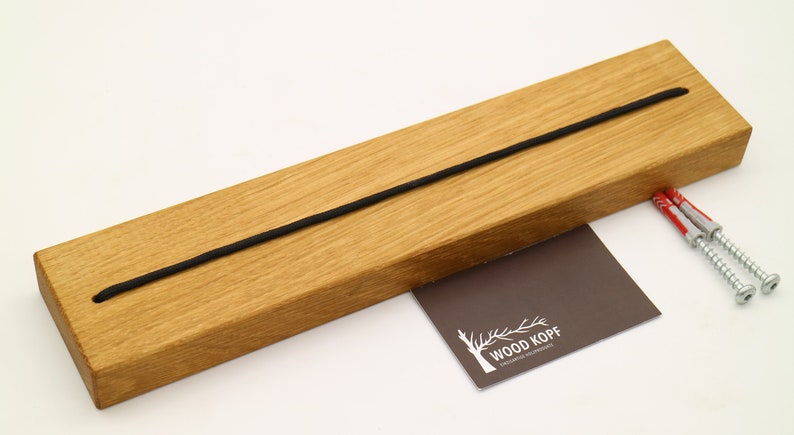 May include: A wooden wall mount with a black elastic cord running through it. The mount is made of light brown wood and has two screw holes on the back. The mount is labeled "WOOD KOPF" on a small brown card.