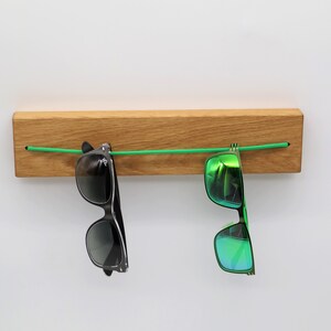 May include: A wooden wall-mounted sunglasses holder with two pairs of sunglasses hanging from a green elastic cord. One pair of sunglasses is black with dark lenses, and the other pair is green with mirrored lenses.