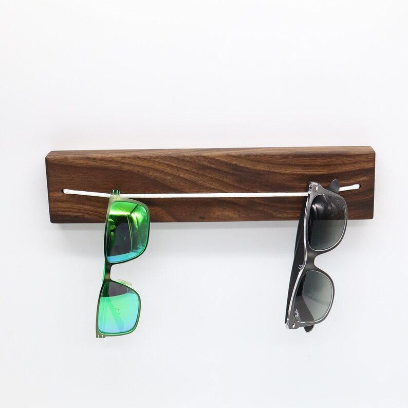 May include: A wooden wall-mounted sunglasses holder with a white cord strung across it. Two pairs of sunglasses are hanging from the cord, one with green lenses and one with black lenses.