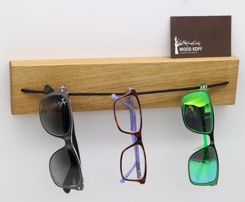 May include: A wooden wall-mounted sunglass holder with a black elastic cord. Three pairs of sunglasses are hanging on the cord. One pair is black, one is green, and one is purple.