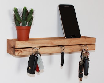 Può includere: Un porta chiavi in legno con uno scaffale in cima. Il porta chiavi ha tre ganci ed è montato su una parete bianca. Una piccola pianta di cactus in vaso è sullo scaffale, e uno smartphone nero è appoggiato sullo scaffale.