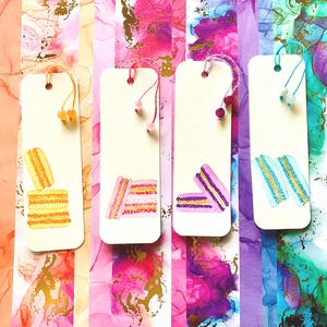May include: Four rectangular paper bookmarks with rounded corners, each featuring a watercolor illustration of two stacked macarons. The bookmarks are accented with colored string loops and small beads in yellow, pink, purple, and light blue.