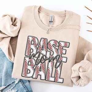 May include: Tan sweatshirt with a baseball-themed graphic that says "BASE mom BALL" in black and red lettering. The graphic is made up of baseballs with red stitching.
