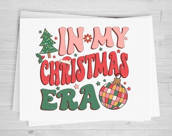 May include: A white card with the text "In My Christmas Era" in a retro font. The text is in pink, red, and green. There is a small Christmas tree, a Santa hat, and a disco ball ornament on the card.