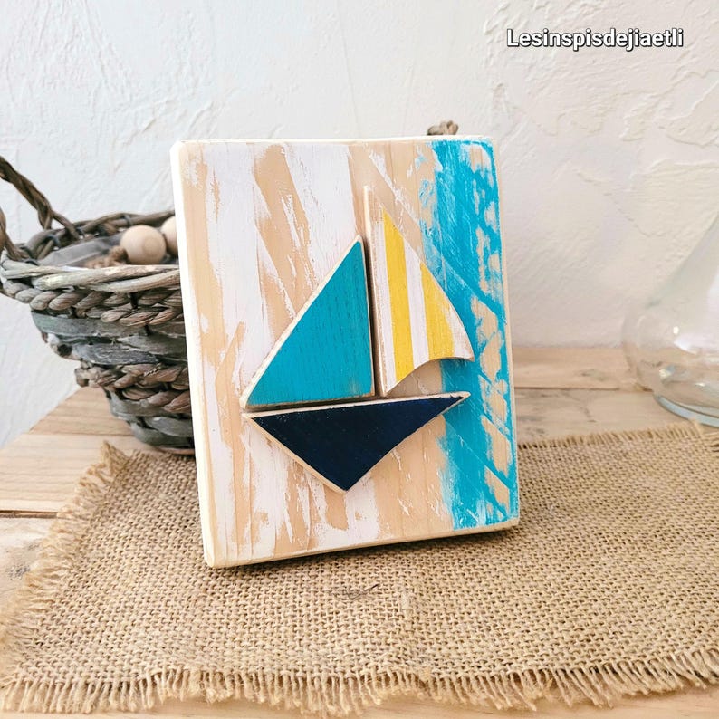 K&ouml;nnte beinhalten: Eine Holz-Wanddekoration mit einem Segelboot aus blau, gelb und wei&szlig; bemaltem Holz. Das Segelboot ist vor einem blau-wei&szlig;en Hintergrund mit einer abgenutzten wei&szlig;en Oberfl&auml;che platziert.
