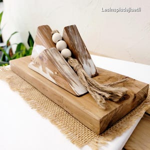Könnte beinhalten: Rustikale Holzdekoration mit drei abgewinkelten Holzelementen mit weißen Farbakzenten, angeordnet auf einer rechteckigen Basis. Drei kugelförmige Perlen und ein Bündel Naturfaserseil ergänzen das Design. Der Text "Lesinspisdejiaetli" ist sichtbar.