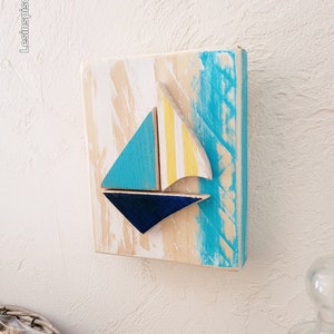 K&ouml;nnte beinhalten: Ein Wandbehang aus Holz mit einem Segelboot-Motiv. Das Segelboot ist aus blauem, gelbem und wei&szlig;em Holz gefertigt. Der Hintergrund ist in Blaut&ouml;nen und Wei&szlig; bemalt.