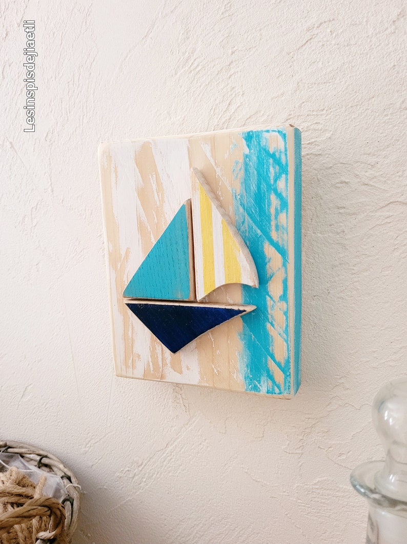 K&ouml;nnte beinhalten: Ein Wandbehang aus Holz mit einem Segelboot-Motiv. Das Segelboot ist aus blauem, gelbem und wei&szlig;em Holz gefertigt. Der Hintergrund ist in Blaut&ouml;nen und Wei&szlig; bemalt.