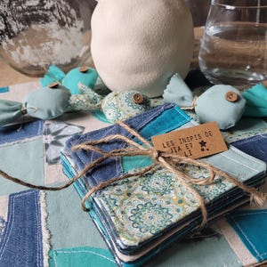 Könnte beinhalten: Ein Stapel quadratischer Untersetzer, mit Bindfaden gebunden, aus Jeansstoff und gemustertem Stoff. Die Untersetzer liegen auf einem Patchwork-Tischset in verschiedenen Blau- und Türkistönen. Das Etikett lautet "LES INSPIS DE JIA ET LI".