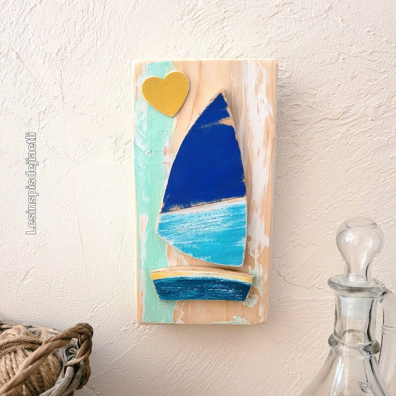 K&ouml;nnte beinhalten: Ein h&ouml;lzerner Wandbehang mit einem blauen und t&uuml;rkisfarbenen Segelboot mit einem gelben Herzen dar&uuml;ber. Der Hintergrund ist in Blaut&ouml;nen, Gr&uuml;n und Wei&szlig; bemalt.