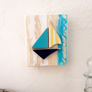 K&ouml;nnte beinhalten: Ein Wandbehang aus Holz mit einem blauen, gelben und wei&szlig;en Segelboot-Design. Das Segelboot besteht aus separaten Holzst&uuml;cken und ist an dem Holzgrund befestigt. Der Hintergrund ist mit einem hellblauen und wei&szlig;en Waschgang bemalt.