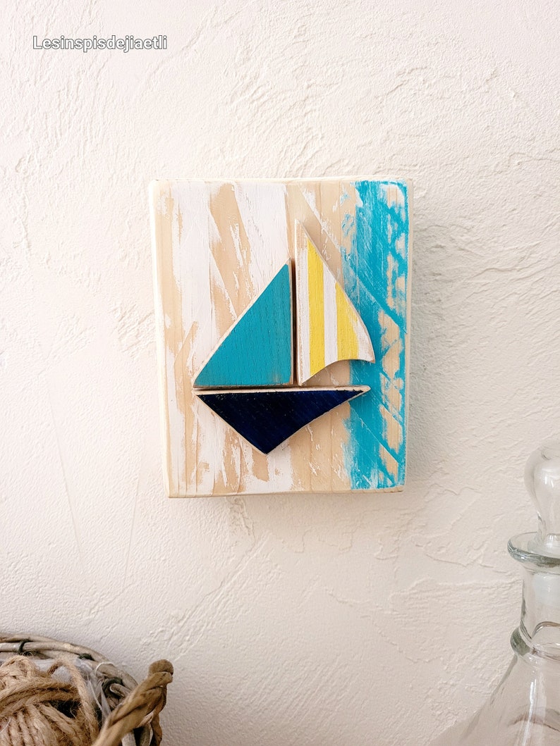 K&ouml;nnte beinhalten: Ein Wandbehang aus Holz mit einem blauen, gelben und wei&szlig;en Segelboot-Design. Das Segelboot besteht aus separaten Holzst&uuml;cken und ist an dem Holzgrund befestigt. Der Hintergrund ist mit einem hellblauen und wei&szlig;en Waschgang bemalt.