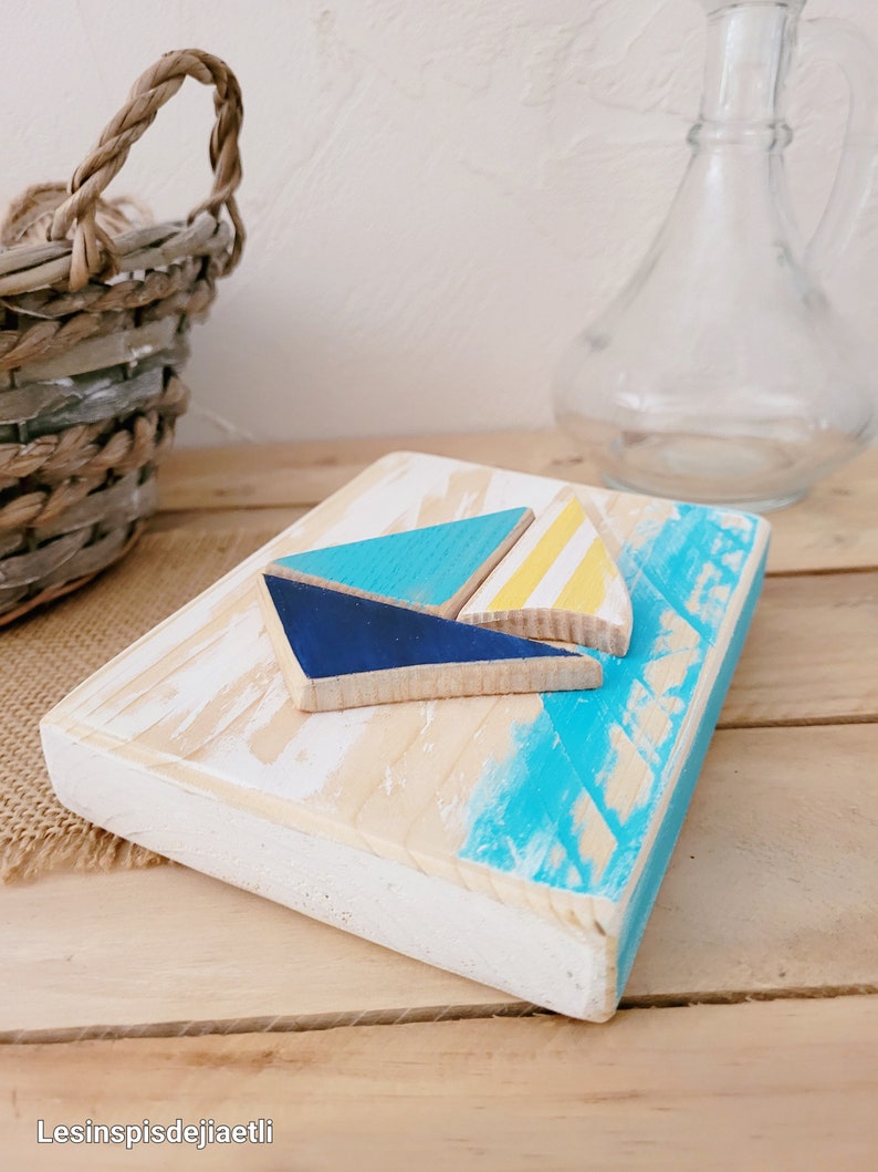 K&ouml;nnte beinhalten: Eine Holz-Wanddekoration mit einem blauen, gelben und wei&szlig;en Segelboot-Design. Das Segelboot besteht aus separaten Holzst&uuml;cken und ist in einem nautischen Thema bemalt.