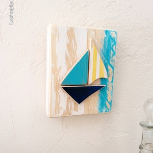 K&ouml;nnte beinhalten: Ein Wandbehang aus Holz mit einem blauen, gelben und wei&szlig;en Segelboot-Design. Das Holz hat eine wei&szlig;e, abgenutzte Lackierung.