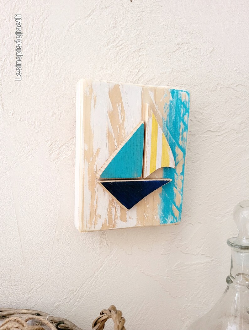 K&ouml;nnte beinhalten: Ein Wandbehang aus Holz mit einem blauen, gelben und wei&szlig;en Segelboot-Design. Das Holz hat eine wei&szlig;e, abgenutzte Lackierung.