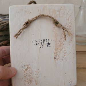 K&ouml;nnte beinhalten: Ein wei&szlig;es Holzschild mit rustikalem Finish. Das Schild hat einen Seilhalter und den Text "LES INSPIS JIA ET LI" mit zwei Sternen.