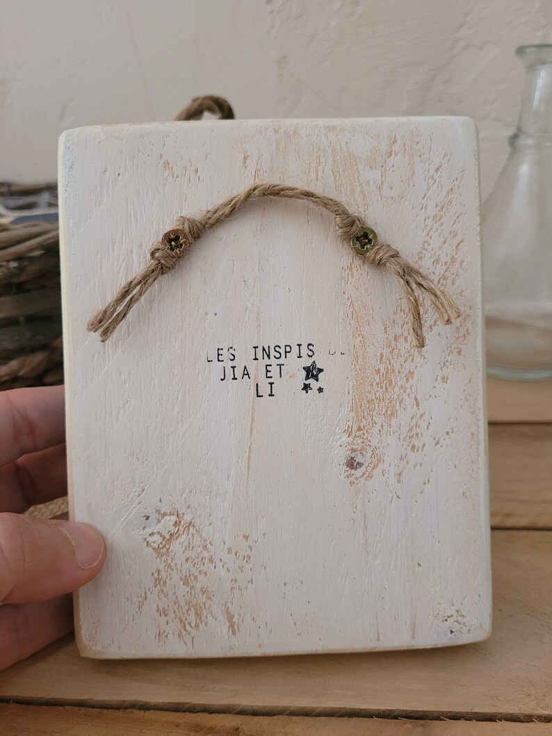 K&ouml;nnte beinhalten: Ein wei&szlig;es Holzschild mit rustikalem Finish. Das Schild hat einen Seilhalter und den Text "LES INSPIS JIA ET LI" mit zwei Sternen.