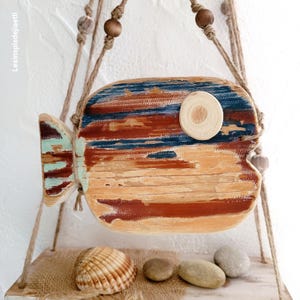 Peut inclure: Un poisson décoratif en bois suspendu avec une peinture vieillie dans des tons bleu, marron et crème. Le poisson possède un œil en bois circulaire et est accroché par une cordelette en jute naturelle avec des perles en bois. Texte visible : Lesinspisdejiaetli.