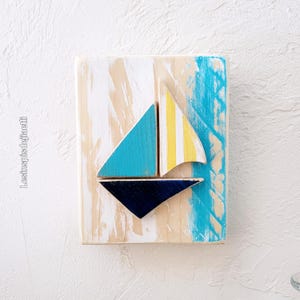 K&ouml;nnte beinhalten: Ein Wandbild aus Holz mit einem Segelboot aus buntem Holz. Das Segelboot ist in Blaut&ouml;nen, Gelb und Wei&szlig; bemalt und vor einem Hintergrund aus wei&szlig; get&uuml;nchtem Holz mit blauen und gelben Akzenten platziert.