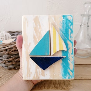 K&ouml;nnte beinhalten: Ein Wandbehang aus Holz mit einem Segelboot-Motiv. Das Segelboot ist aus blau, gelb und wei&szlig; bemaltem Holz gefertigt. Der Hintergrund ist in Blaut&ouml;nen und Wei&szlig; bemalt.