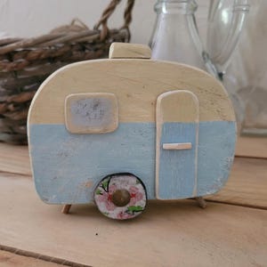 Könnte beinhalten: Ein kleiner Wohnwagen aus Holz, bemalt in Hellblau und Creme. Der Wohnwagen hat ein kleines Fenster, eine Tür und ein dekoratives Rad mit floralem Design. Der Wohnwagen steht auf einer Holzoberfläche. Der Text "Lesinspisdejiaetli" steht unten.