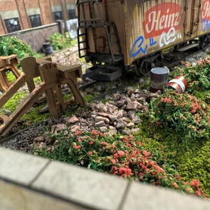 May include: A miniature model of a weathered freight train car with the Heinz logo and graffiti. The car is brown with rust and sits on a track surrounded by miniature plants, rocks, and a brick building in the background.