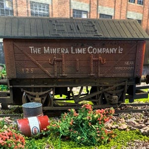May include: A detailed model of a vintage brown freight train car with the text "The Minera Lime Company Ld" on the side. The model includes wheels, a roof, and doors. A small red and white Esso barrel and a metal can are in front of the train car.