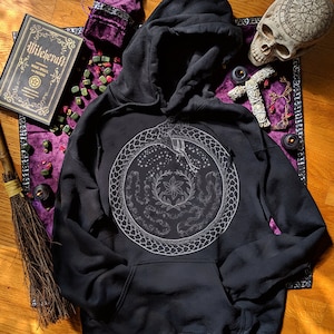 May include: Black hooded sweatshirt with a white graphic print of a snake wrapped around a circle with stars, flowers, and vines. The text "Witchcraft" is visible on a book in the background.
