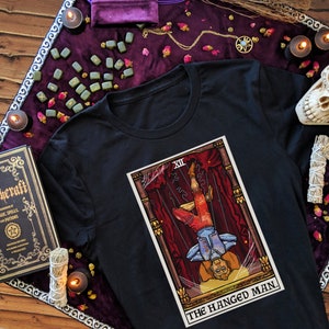 Puede incluir: Camiseta negra con un gráfico de la carta del tarot El Ahorcado. La carta está ilustrada en un estilo vintage con colores rojo, amarillo y azul. El número de la carta XII es visible en la esquina superior izquierda. El texto "THE HANGED MAN" está impreso debajo de la carta.