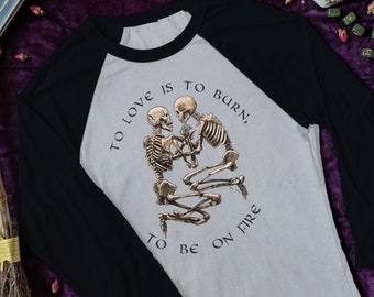 May include: A beige and black baseball tee shirt with a graphic of two skeletons holding hands. The text on the shirt reads "To love is to burn, to be on fire".